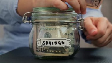 Tanınmayan kadın, para biriktirme kavanozuna dolar banknotlarıyla dolu para ekliyor. Tasarruf metni kavanozun önünde. Gelecekteki güvensizlik için kişisel finansman yöneticiliği