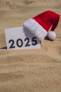 2025 yazısında arka plandaki kumlu sahil güneşinde tebrik kartı var. Noel Baba şapkası Yeni Yıl Dekorasyonu. Yaz tatili dekoru. Tatil konsepti takvim tarihi kartpostalı. Uzaklaşıyorum Seyahat ediyorum