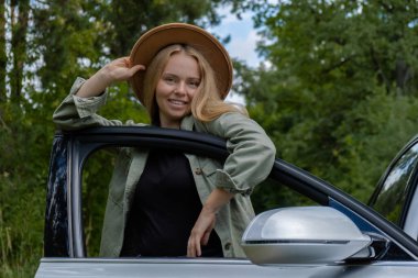 Smiling young woman in hat stoped on road to make a break in driving car. Local solo travel on weekends concept. Exited woman explore freedom outdoors in forest. Unity with nature lifestyle, rest clipart