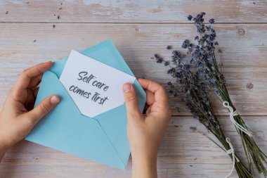 Female hands taking paper card note with text SELF CARE COMES FIRST from blue envelope. Lavender flower. Top view, flat lay. Concept of mental spiritual health self care wellbeing mindfulness clipart