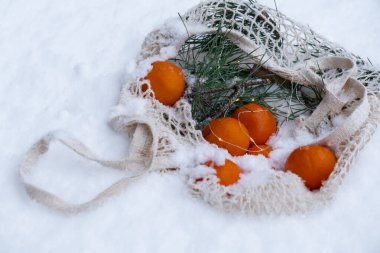 Karlı kışta dışarıda köknar dalıyla birlikte yeniden kullanılabilir ağ torbasında olgun mandallar. Sürdürülebilir Noel kutlaması, sorumluluk ve kapsayıcılık kavramı. Pamuk torbadaki portakallar