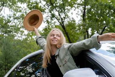 Arabanın camından bakan şapkalı, gülümseyen genç bir kadın. Hafta sonları yerel solo seyahat. Çıkan kadın ormanda özgürlüğü keşfediyor. Doğa yaşam tarzıyla birlik, dinlenme rahatlama 