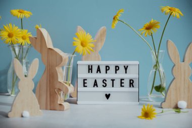 Phrase HAPPY EASTER traditional greeting to christian holiday. Festive mood composition with bunnies and yellow flowers on pastel blue background. Sustainable decorations clipart