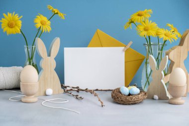 Boş beyaz zarf, şenlikli mavi arka plan ve çevre dostu sıfır atık doğal dekorasyonda kartpostal fotokopi alanı oluşturur. Sarı papatya çiçekleri. Tavşan yumurtası. Tatil tebrik kartı.