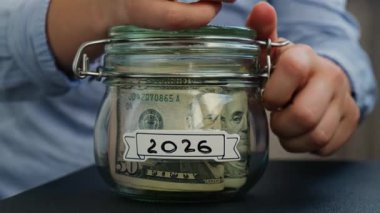 Kadın eli para ekler. 2026 yılına ait bir kavanoz dolusu Amerikan doları nakit para. Hazırlık para tasarrufu. Orta karar tüketim ve ekonomi. Para toplama
