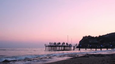 Günbatımında Antalya Türkiye 'deki ahşap iskeleye dalgalar çarpıyor. Palmiyeler ve deniz seyahati turizmi konsepti. Kumsal ve dağlar. Huzur ve yavaşlama 
