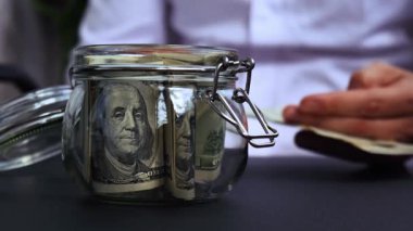 Adam kumbaradaki cam kavanozdan gelen dolar banknotlarını sayıp sayıyor. Tanınmayan bir iş adamının eli kulağında. Bütçe yatırımı kavramını sakla. Amerikan doları.