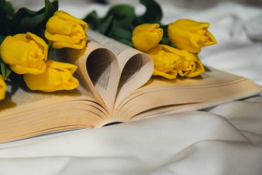 Kalp sembolü şeklinde kitap sayfaları dağınık yatakta pembe lale buketi ile aşk. Romantik sabah sahnesi. Tebrik kartı. Bahar zamanı çiçekleri.