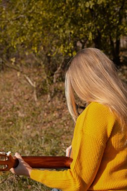 Güzel sarışın kız akustik gitar çalıyor ve şarkı söylüyor güneşli bir sonbahar gününde dışarıda eğleniyor. Hippi kadın boho stilinde öğreniyor ve müzik enstrümanı çalıyor. Sonbahar yaprakları