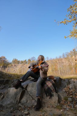 Güzel sarışın kız akustik gitar çalıyor ve şarkı söylüyor güneşli bir sonbahar gününde dışarıda eğleniyor. Hippi kadın boho stilinde öğreniyor ve müzik enstrümanı çalıyor. Sonbahar yaprakları