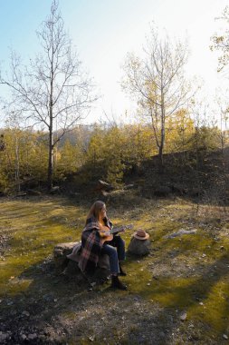 Güzel sarışın kız akustik gitar çalıyor ve şarkı söylüyor güneşli bir sonbahar gününde dışarıda eğleniyor. Hippi kadın boho stilinde öğreniyor ve müzik enstrümanı çalıyor. Sonbahar yaprakları