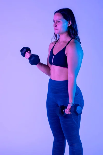 stock image Young latin woman exercising with dumbbells, standing, in studio with color background