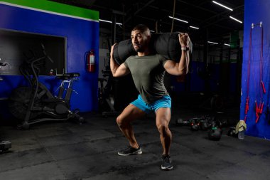 Spor giyimli bir adam spor salonunda sırtında kum torbasıyla çömeliyor.