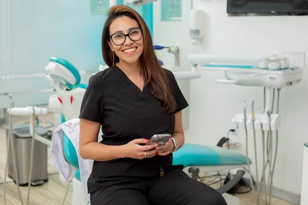 年轻的女牙医笑着看着摄像机 手里拿着手机坐在办公室里 — 图库照片