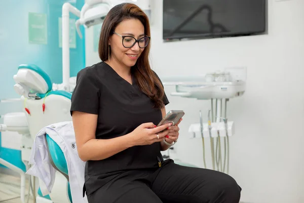 Young Latina dentist sitting in her office, checking her cell phone