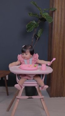 Küçük kız yemek sandalyesinde elinde kaşıkla oturmuş tabağına yemek ile bakıyor. Bebek sütten kesme konsepti