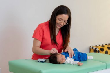 Gülümseyen çocuk doktoru tıbbi muayene masasında yatan yeni doğmuş bir bebeğin işitme tepkilerini ölçmek için bir cihaz kullanıyor.