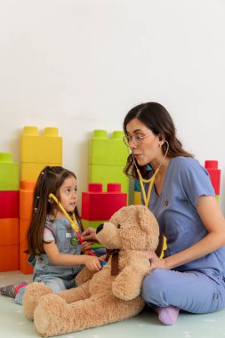 Pediyatrist ve küçük kız stetoskop kullanıyor, oyuncak ayıyla doktorculuk oynuyor, sağlık ve çocukluk gelişimini destekliyor.