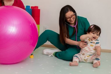 Genç bayan pediatrist steteskopla neşeli bir bebeği inceliyor ve pozitif bir sağlık deneyimini destekliyor.