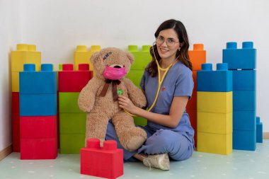 Genç bayan pediyatrist steteskop kullanıyor, oyuncak ayıyla yüz maskesi takıyor, klinikte renkli oyuncak bloklarıyla oturuyor.