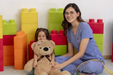 Neşeli bir oyun odasında gülümseyen bir çocuk doktoru oyuncak ayı tutan genç bir kızla etkileşime girer ve dost canlısı bir sağlık hizmeti ortamı sağlar.