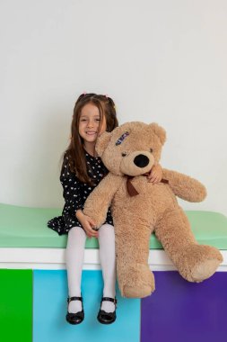 Genç bir kız çocuk doktorunun ofisinde büyük bir oyuncak ayıyla oturup neşeli ve rahatlatıcı bir sağlık hizmeti sunuyor.