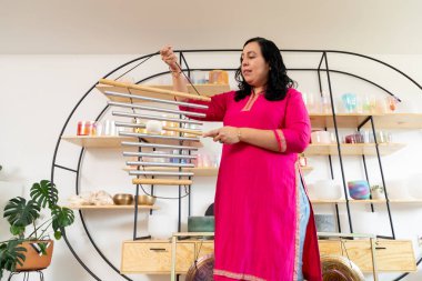 Bir müzik stüdyosunda rüzgar çanları çalan Latin bir kadın ses iyileştirme konusunda uzmanlaşmış, etrafında Tibet kâseleri, gonglar ve pervaneler var.