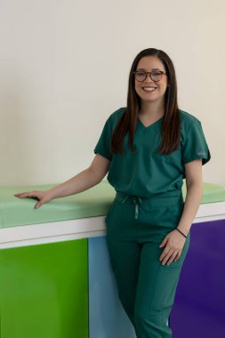 Renkli ofisinde muayene masasının yanında dururken gülümseyen kendinden emin genç bir çocuk doktoru.