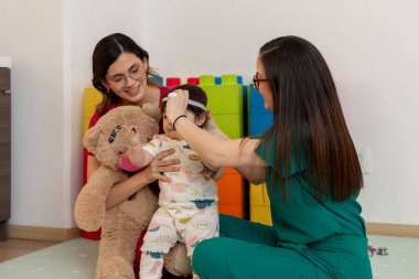 Pediyatrist, rutin kontroller sırasında bebeğin kafasının büyümesini ölçmek için mezura kullanıyor. Annesi onu ve oyuncak ayıyı tutuyor.