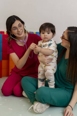 Neşeli bir muayenehanede steteskopla neşeli bir bebeği muayene eden iki kadın pediyatrist.