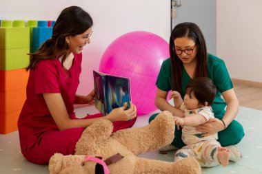 İki gülümseyen çocuk doktoru kitap okuyarak ve muayenehanede oyuncak ayıyla oynayarak bir kızı eğlendiriyorlar.