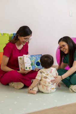 İki gülümseyen çocuk doktoru, tıp bürosunda oyun hasırında oturan bir bebeğe renkli bir kitap okuyor.