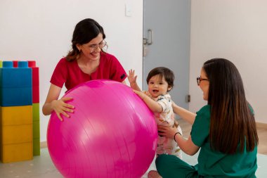İki gülümseyen kadın pediyatrist, tıbbi muayenehanede terapi seansı sırasında bebeğin büyük pembe topuyla oynamasına yardım ediyor.