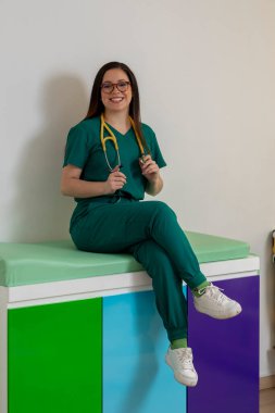 Elinde steteskopla gülümseyen ve renkli muayenehanesinde oturan genç bir çocuk doktoru.