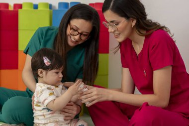 İki gülümseyen kadın pediyatrist renkli ofiste neşeli terapi seansı sırasında küçük kızla etkileşime geçiyorlar.