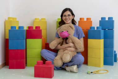 Genç bir çocuk doktoru, ofisinde yerde otururken oyuncak ayıya sarılıyor.