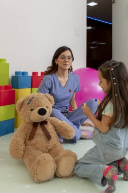 Küçük bir kız ve oyuncak ayıyla etkileşime giren genç bir çocuk doktoru doktorun ofisinde rahat ve neşeli bir atmosfer yaratıyor.