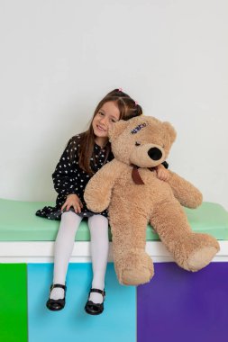 Genç hasta oyuncak ayısını kucaklıyor, renkli bir sağlık ortamında iç açıcı bir rahatlık ve bakım sahnesi yaratıyor.