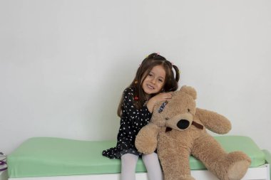 Çocuk doktorunun muayenehanesinde muayene masasında otururken oyuncak ayısına sarılan küçük kız güven ve rahatlık gösteriyordu.