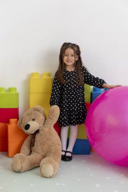 Geniş bir oyuncak ayı ve şişme pembe bir topun yanında duran genç kız, çocuk sağlığı ve refahı için çocuk sağlığı ve pediyatristin ofisinde renkli yapı taşlarıyla çevrili.