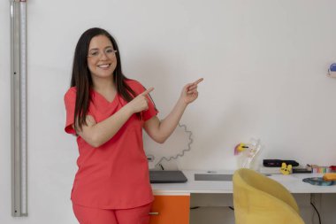 Meksikalı bayan çocuk doktoru gülümseyip renkli ofisinde her iki tarafı işaret ederek genç hastalar için hoş bir ortam yaratıyor.