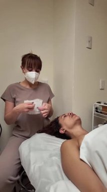 Profesyonel güzellik uzmanı, güzellik merkezinde masaj yatağında yatan bir müşterinin yüzüne canlandırıcı bir maske takıyor.