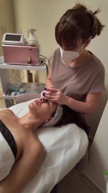 Maske takan, yüz kremi süren ve güzellik salonundaki masaj masasında yatan müşteriye boyun masajı yapan profesyonel bir güzellik uzmanı.