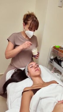 Profesyonel güzellik uzmanı, bir sağlık merkezinde masaj masasında yatan genç bir kadının yüzüne nemlendirici maske takıyor.
