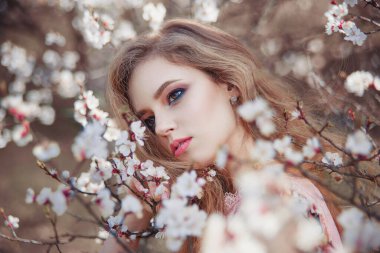 Baharda çiçek açan bir bahçede uzun saçlı güzel bir kızın portresi. Doğanın tadını çıkarıyor. Güzel makyajlı sağlıklı bir kadın. Bahar konsepti Bahçedeki güzel kız. Etrafta kayısı ağacı çiçekleri var. Arkaplan bulanık