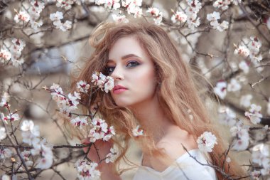 Baharda çiçek açan bir bahçede uzun saçlı güzel bir kızın portresi. Doğanın tadını çıkarıyor. Güzel makyajlı sağlıklı bir kadın. Bahar konsepti Bahçedeki güzel kız. Etrafta kayısı ağacı çiçekleri var. Arkaplan bulanık