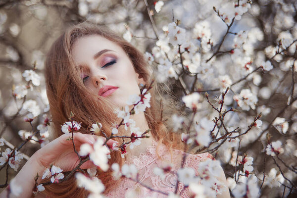 Portrait of a beautiful girl with long hair in a blooming garden in spring. Enjoying nature with closed eyes. Healthy woman outdoors. Spring concept. Beautiful girl in the garden. There are apricot tree flowers around. The background is blurry