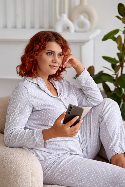 Pijamalı bir kadın elinde telefonla bir sandalyede oturuyor. Ev ortamı