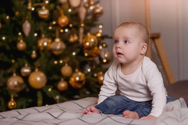 Bebek Noel ağacının önünde oturuyor ve sola bakıyor. Tatili bekliyorum.