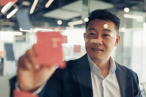 Asian Businessman writing on sticky notes on glass wall while working in modern office